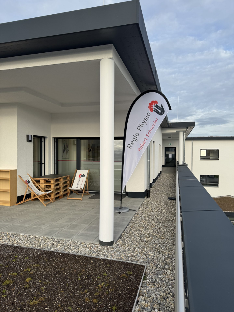 Physiotherapie-Raum mit hellgrüner Behandlungsliege, Kettlebells, Hanteln und großem Fenster mit Blick auf grüne Hügel.